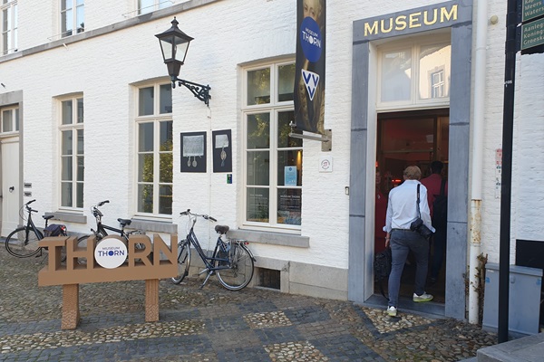 Ingang van Museum Thorn, gevestigd in een wit historisch pand met een open deur waar bezoekers naar binnen gaan. Voor het gebouw staat een houten bord met de tekst "THORN" en een logo. Enkele fietsen staan geparkeerd tegen de muur, en een lantaarn hangt aan de gevel.