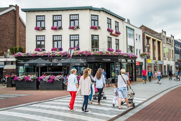 Shoppers cross in the centre of Weert, passing a terrace with flowers and several shops in the background.
