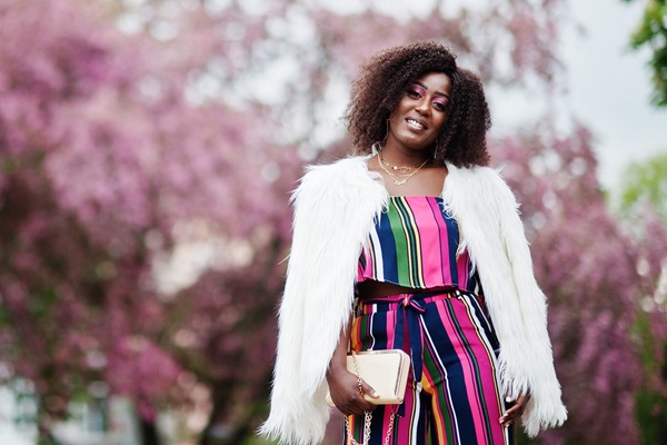 Vrouw met een kleurrijke jumpsuit en witte fluffy jas poseert lachend voor bloeiende roze bomen in de lente.