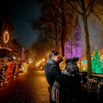 Twee volwassenen in warme winterjassen en mutsen staan dicht bij elkaar en kijken lachend omhoog terwijl sneeuwvlokken vallen op het sfeervol verlichte Munsterplein in Roermond.