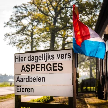 Bord bij een boerderijwinkel met verse asperges, aardbeien en eieren, langs een landelijke weg in Limburg.